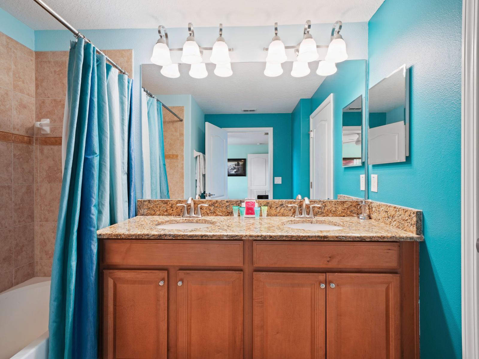 - Vibrant bathroom with turquoise walls, featuring a double vanity with granite countertops and wood cabinetry - Tiled bathtub with a blue curtain adds a splash of color and texture - Bright lighting above the mirror enhances the fresh, modern feel