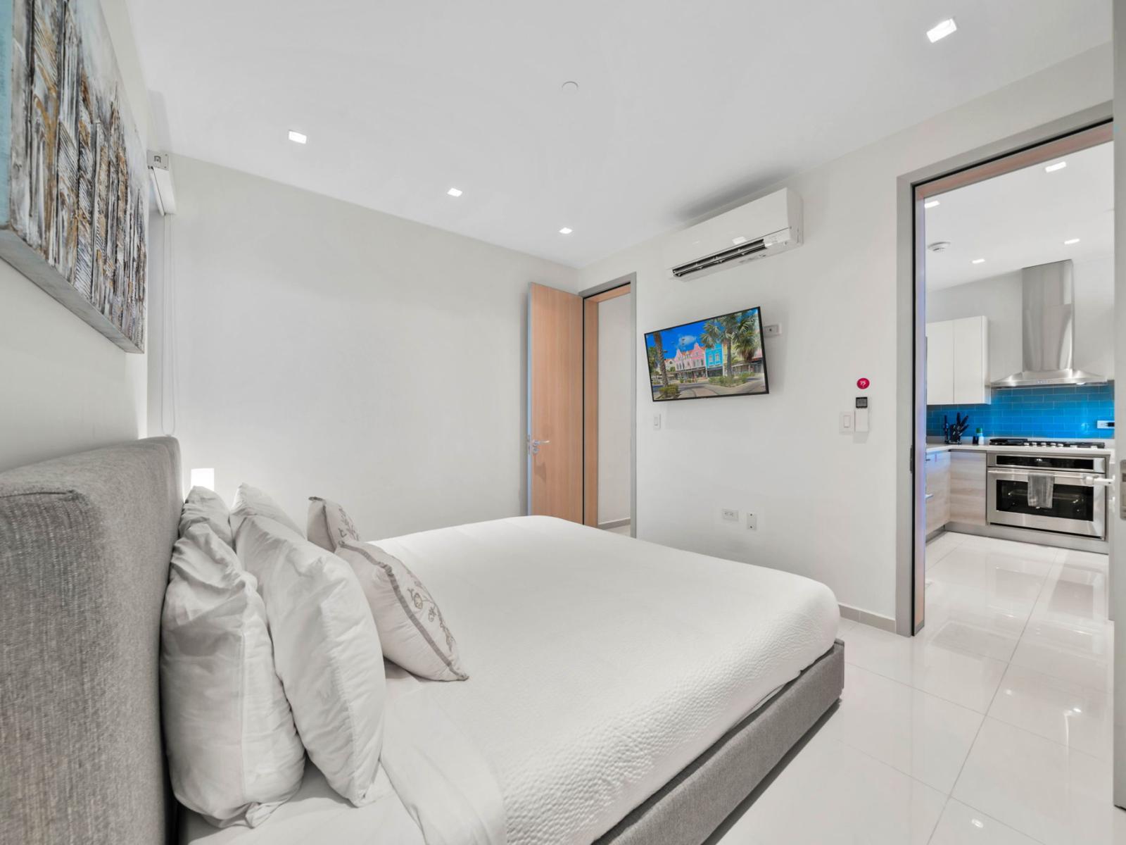 - Alluring bedroom featuring a king-sized bed with soft linens and graceful decor - Wall-mounted TV provides entertainment, while the door opens to the kitchen - The clean, elegant design creates a serene and inviting atmosphere for rest