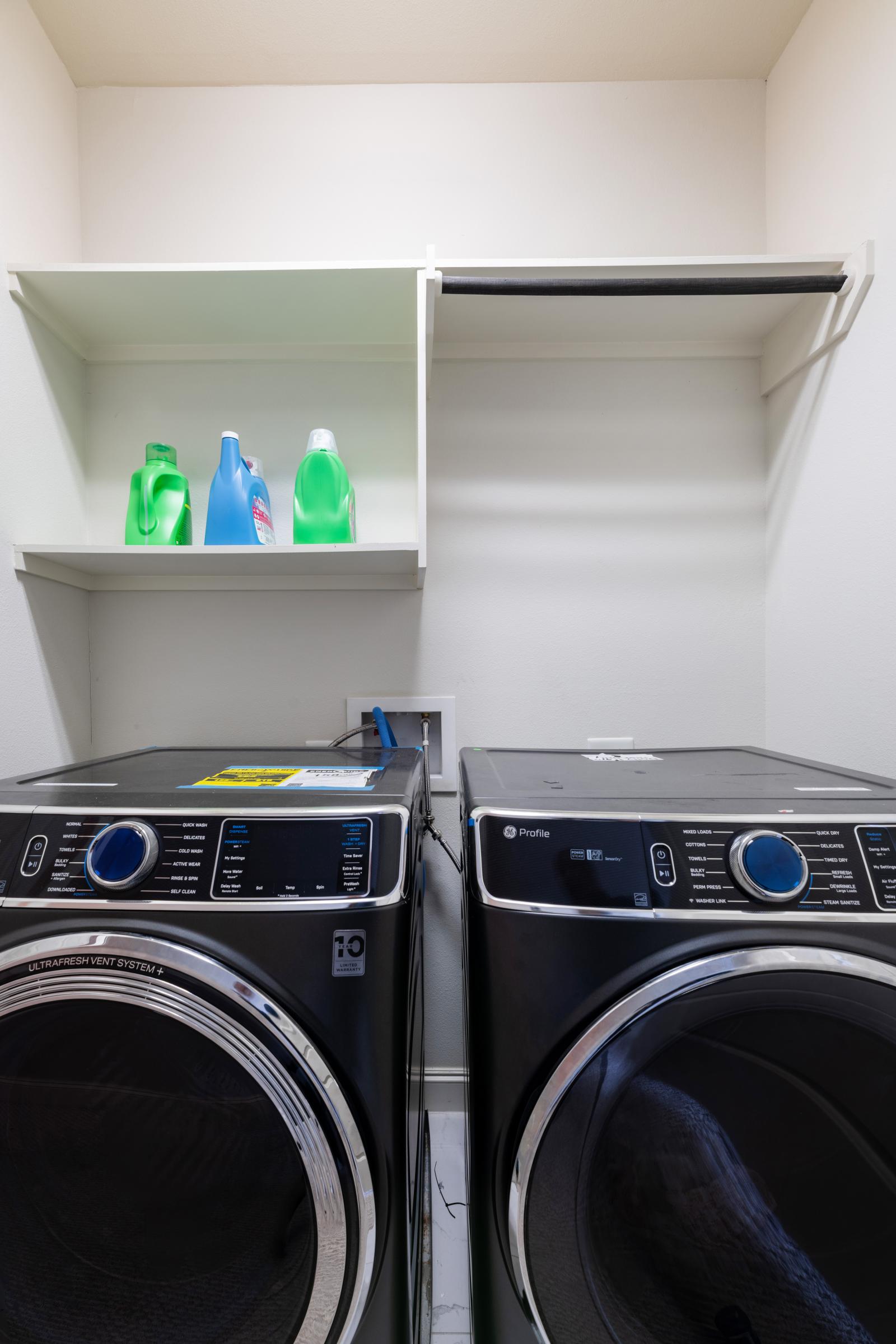 A sleek laundry area featuring high-efficiency GE Profile washer and dryer units, ample overhead shelving for detergents, and a built-in hanging rod for added convenience.