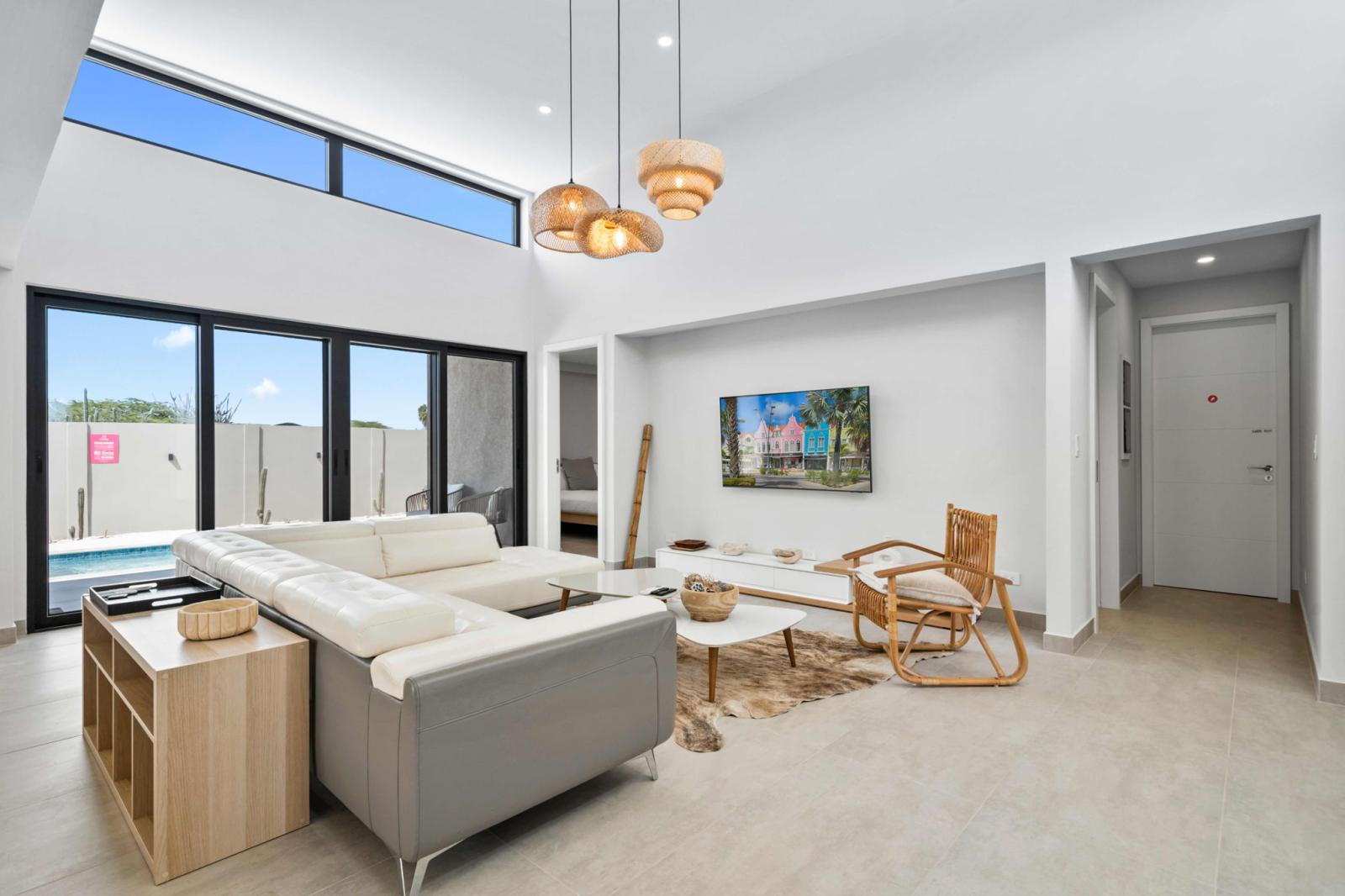 Bright & inviting living space with floor to ceiling windows leading to the pool - Modern design with cozy seating, stylish lighting & a mounted smart TV for entertainment - Perfect for relaxing after a beach day, with seamless indoor outdoor flow