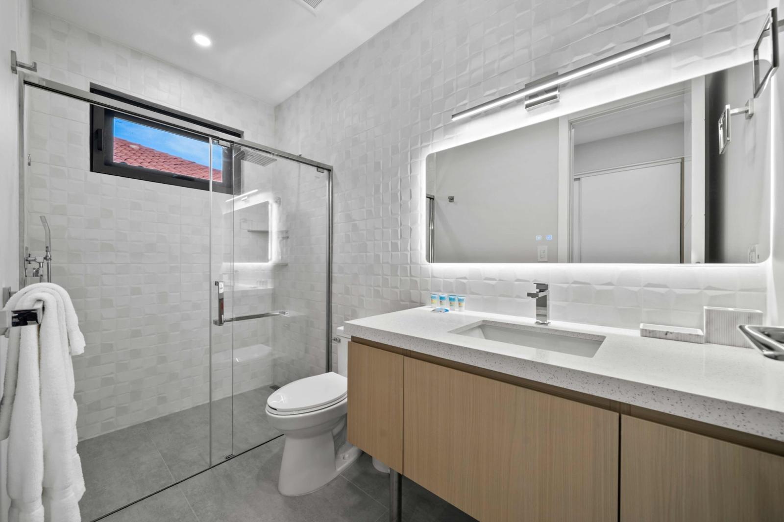 Modern bathroom featuring a sleek vanity, LED mirror, and glass-enclosed shower - Fresh towels and essential toiletries provided for a refreshing and hassle free stay - Designed for comfort with clean lines, soft lighting, and a spa like atmosphere