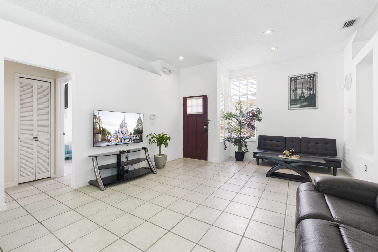 Welcoming front living area featuring neutral tile flooring, a mounted smart TV, and large windows that fill the space with natural light—perfect for relaxing after a day at nearby attractions.