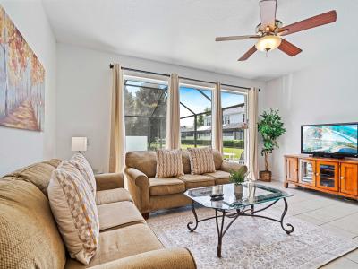 Cozy and bright living area with large windows providing a view of the private pool - Comfortable seating with plush sofas and a table perfect for family gatherings - Ceiling fan and TV for entertainment, creating a relaxing and enjoyable space