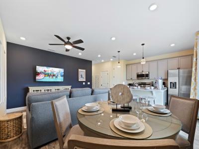 Cozy open-concept dining area with navy accent wall and mounted TV - Dining table set for four, pendant lights over a modern kitchen with stainless steel appliances - Natural light from large windows with patterned curtains brightens the space