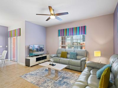 Cozy living room featuring plush leather sofas in soft gray tones, accented with colorful pillows - Striped window valances add a playful touch, complementing the light purple walls - Ceiling fan and modern decor create a welcoming atmosphere